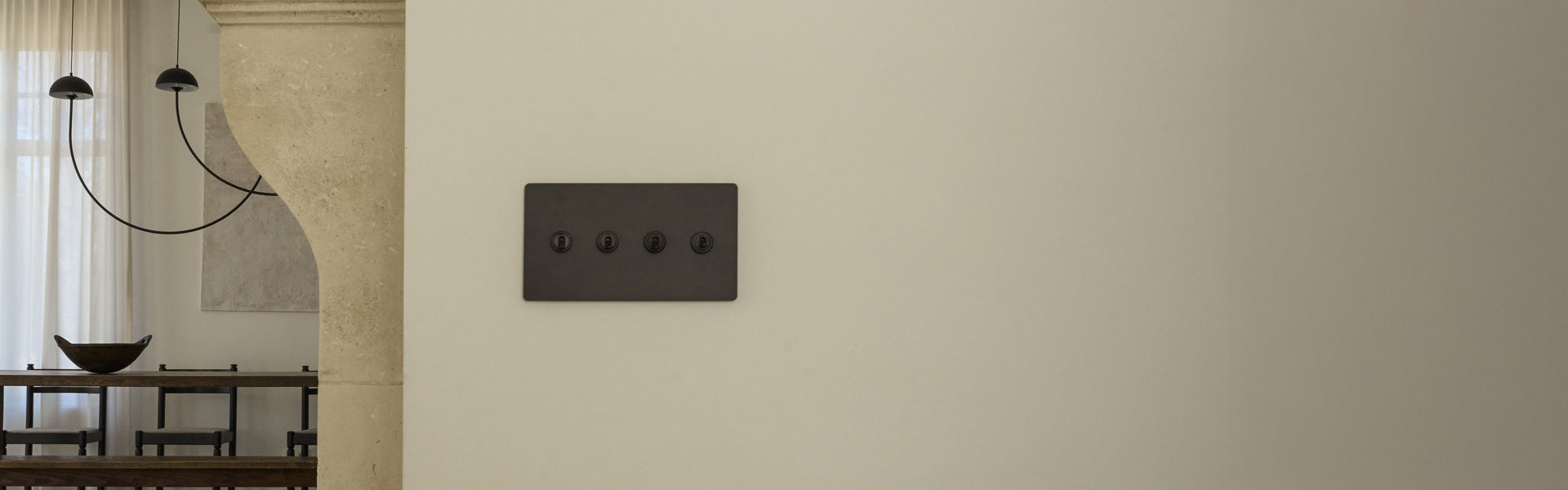 A minimalist light switch panel with four black switches on a cream wall, adjacent to a rustic dining area with a chandelier.