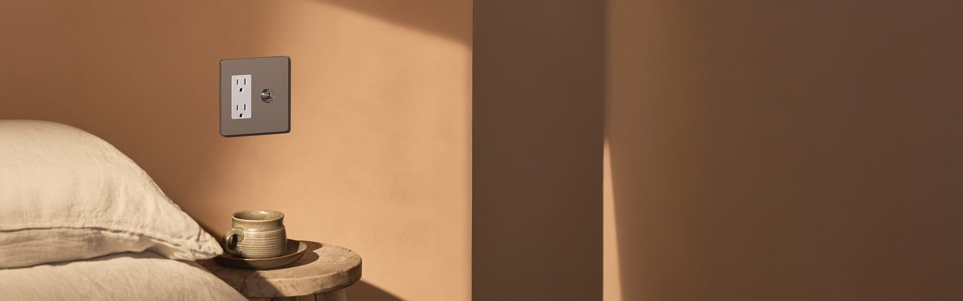 A minimalist bedside scene featuring a clay cup on a wooden table, with a wall switch above a peach-colored wall. Soft bedding is visible.
