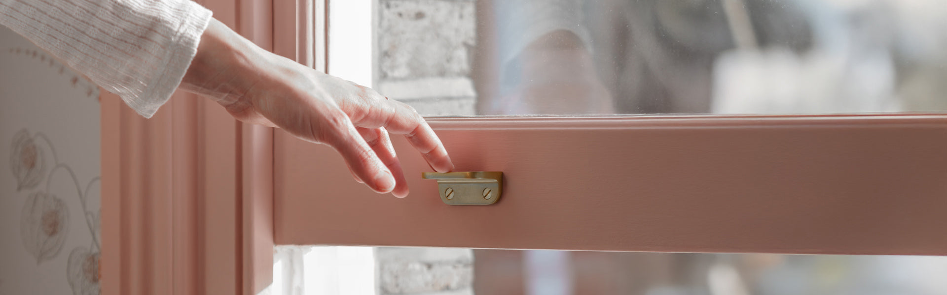 A hand reaching to unlock a brass window latch on a pink window frame, sunlight streaming through the glass.