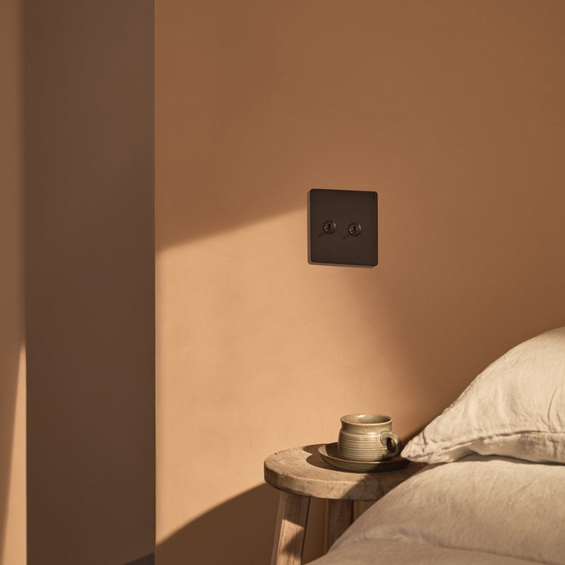 A modern black power outlet on a soft beige wall, with a wooden stool beside a plush bed and a cup resting on the stool.