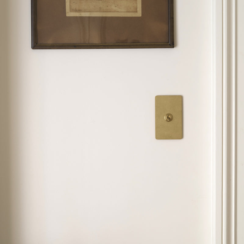 A vintage landscape artwork framed in black hangs on a white wall beside a brass wall plate, with a wooden chair partially visible.