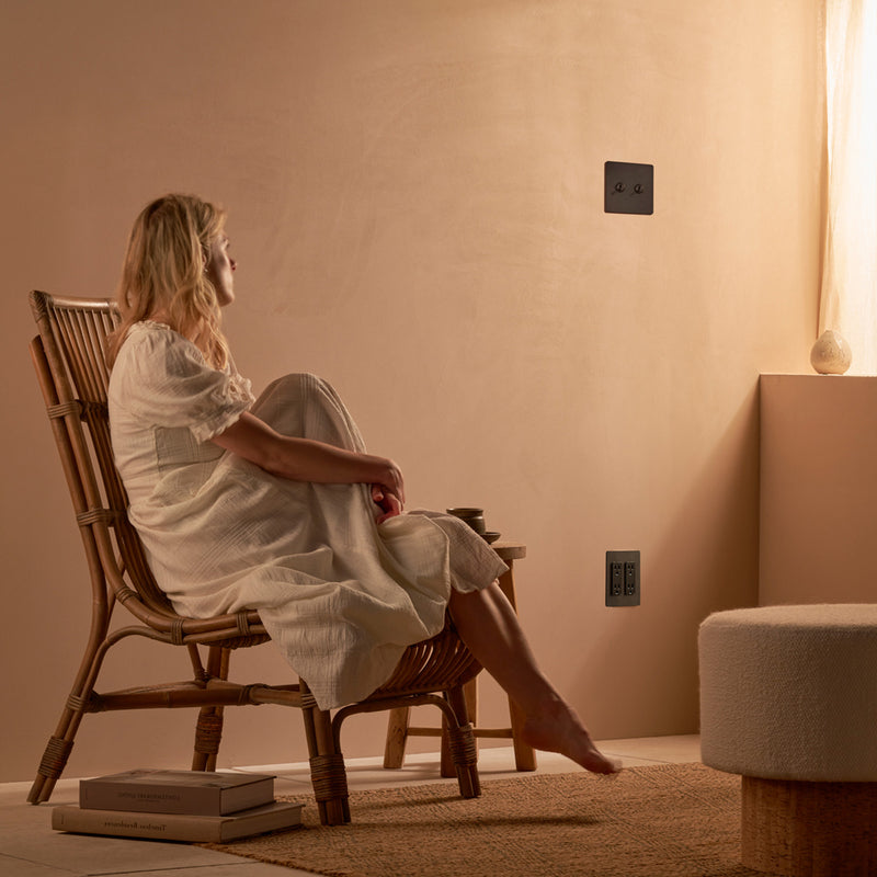 A woman in a white dress sits in a wicker chair, gazing thoughtfully at a minimalist, light-colored wall with a sleek black outlet.