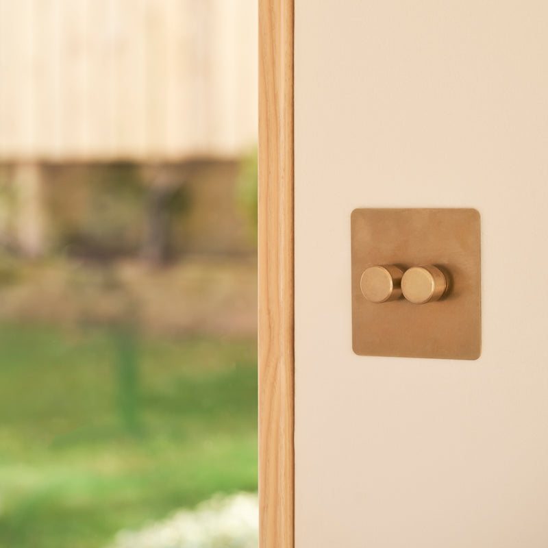 Close-up of a stylish brass light switch mounted on a cream wall, with a blurred garden background featuring greenery and flowers.