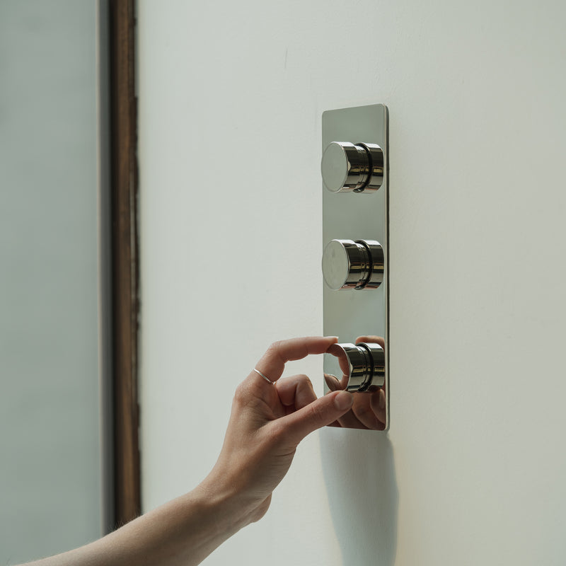 A hand adjusting a sleek, modern metallic knob on a minimalist wall panel with three circular controls.