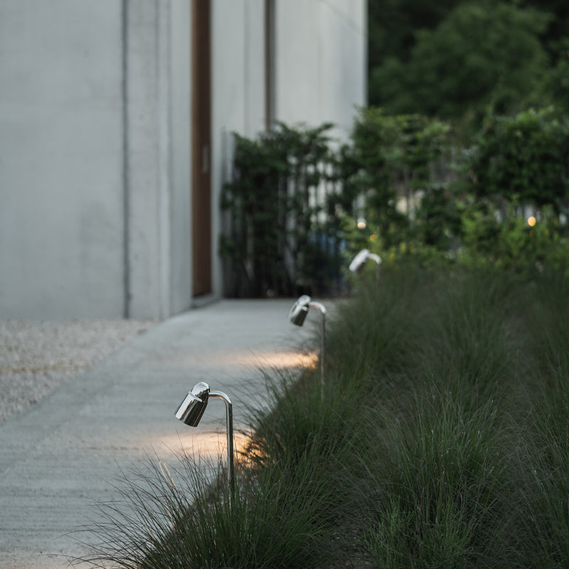 A modern pathway lined with sleek outdoor lights, leading to a minimalistic concrete building surrounded by lush greenery.