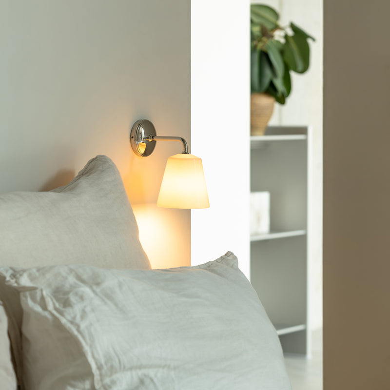 Cozy bedroom corner featuring a soft pillow, wall-mounted lamp, and greenery in a light, modern space.