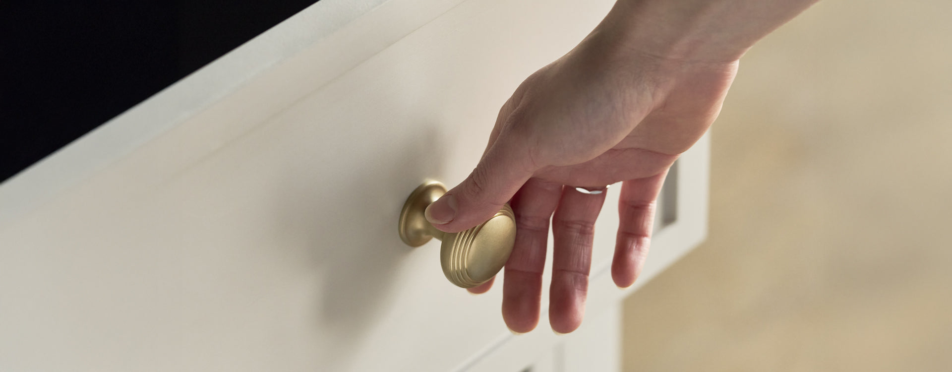 A hand reaching for a brass cabinet knob on a white drawer, ready to open it.
