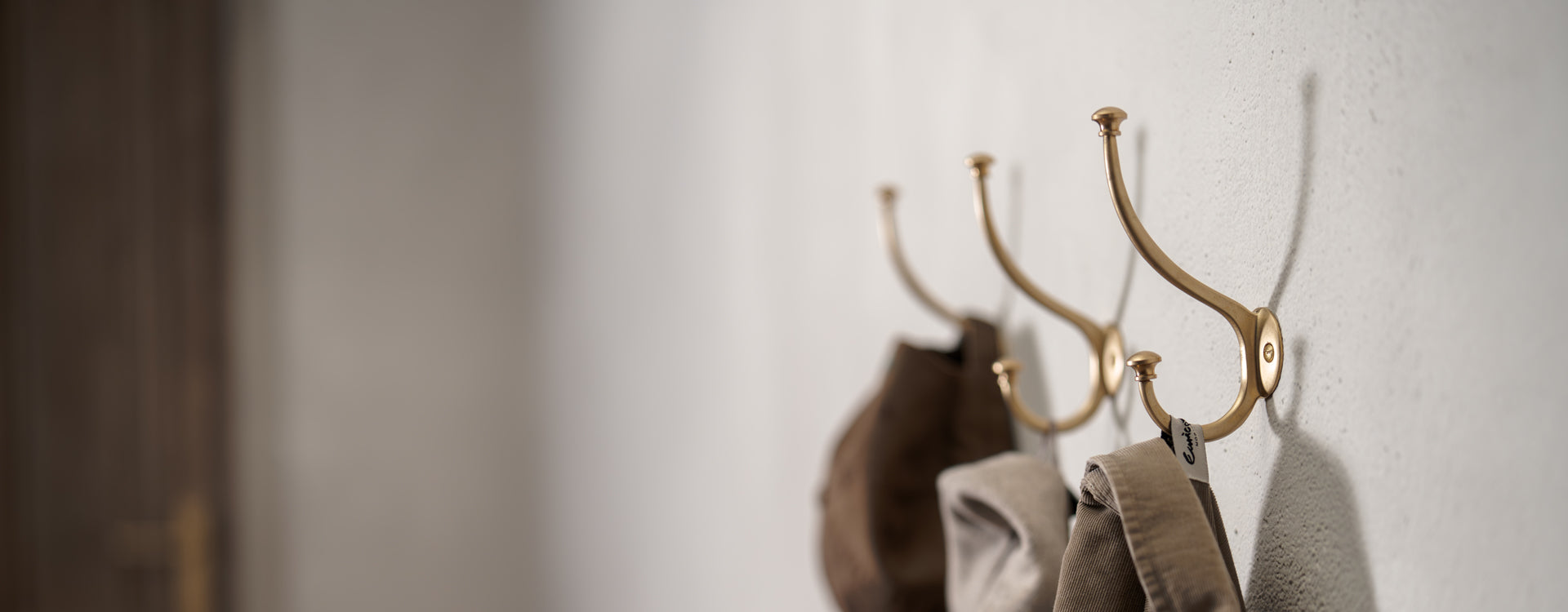 Two elegant brass wall hooks support soft fabric hats against a textured white wall, showcasing a minimalist design.
