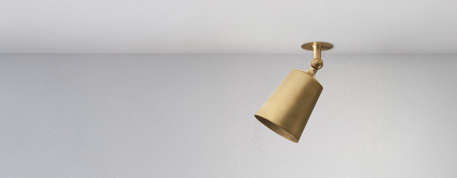 A sleek, modern brass pendant light hangs from a ceiling, casting a warm glow against a plain white wall.
