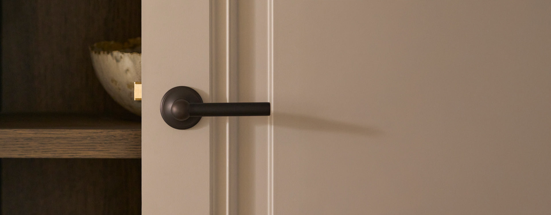 Close-up of a sleek black door handle on a light-colored door, with a decorative bowl visible on a wooden shelf behind it.
