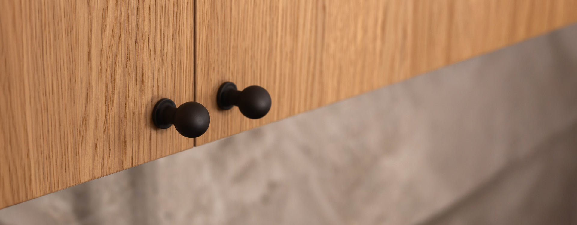 Close-up of two black round cabinet knobs on light wooden cabinet doors with a subtle grain texture.
