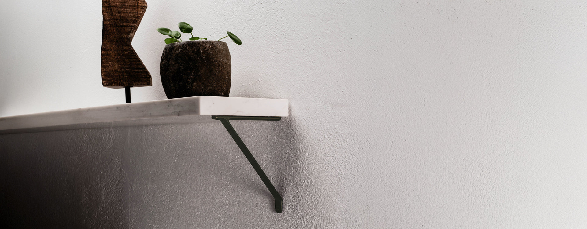 A minimalist shelf with a marble top, supported by a green bracket, displays a dark pot plant and a wooden sculpture against a white wall.
