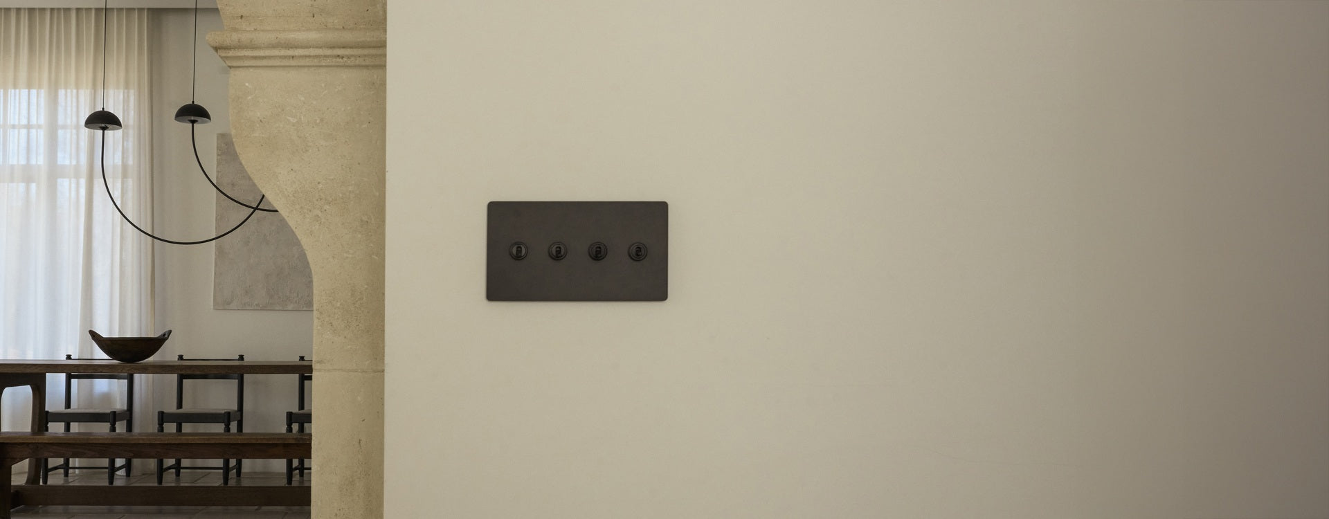 A minimalist light switch panel with four black switches on a cream wall, adjacent to a rustic dining area with a chandelier.
