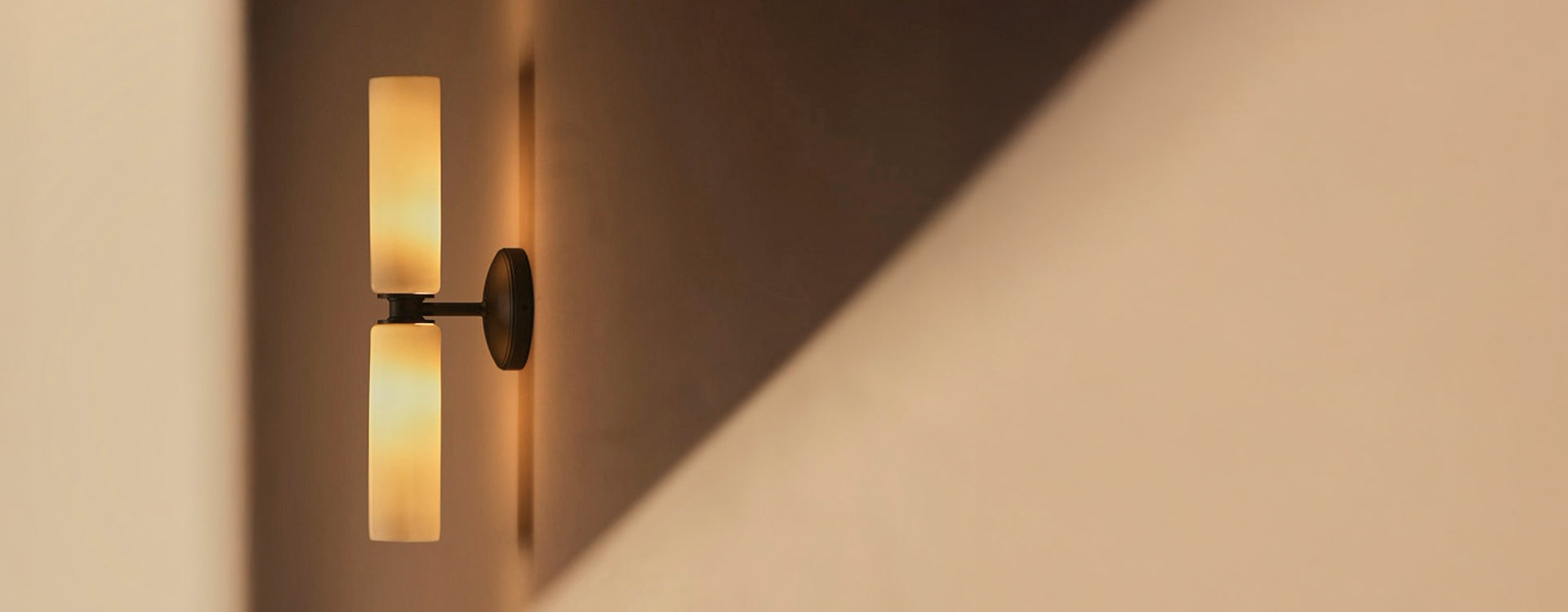 Wall-mounted lamp with two cylindrical frosted glass shades casting a warm glow, creating soft shadows on a beige wall.
