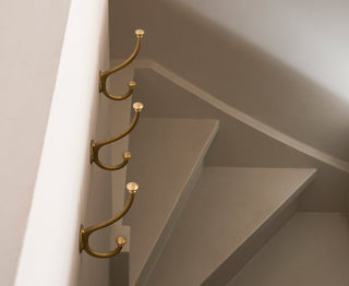 Four elegant brass hooks mounted on a wall beside a staircase, modern design against a neutral backdrop.
