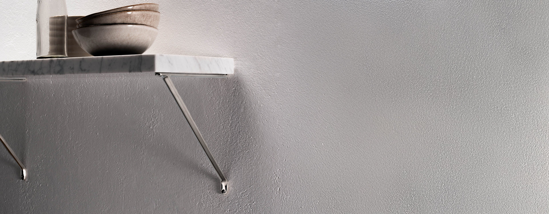 A marble shelf supported by a sleek metallic bracket, with two ceramic bowls and a glass bottle resting on top against a textured wall.
