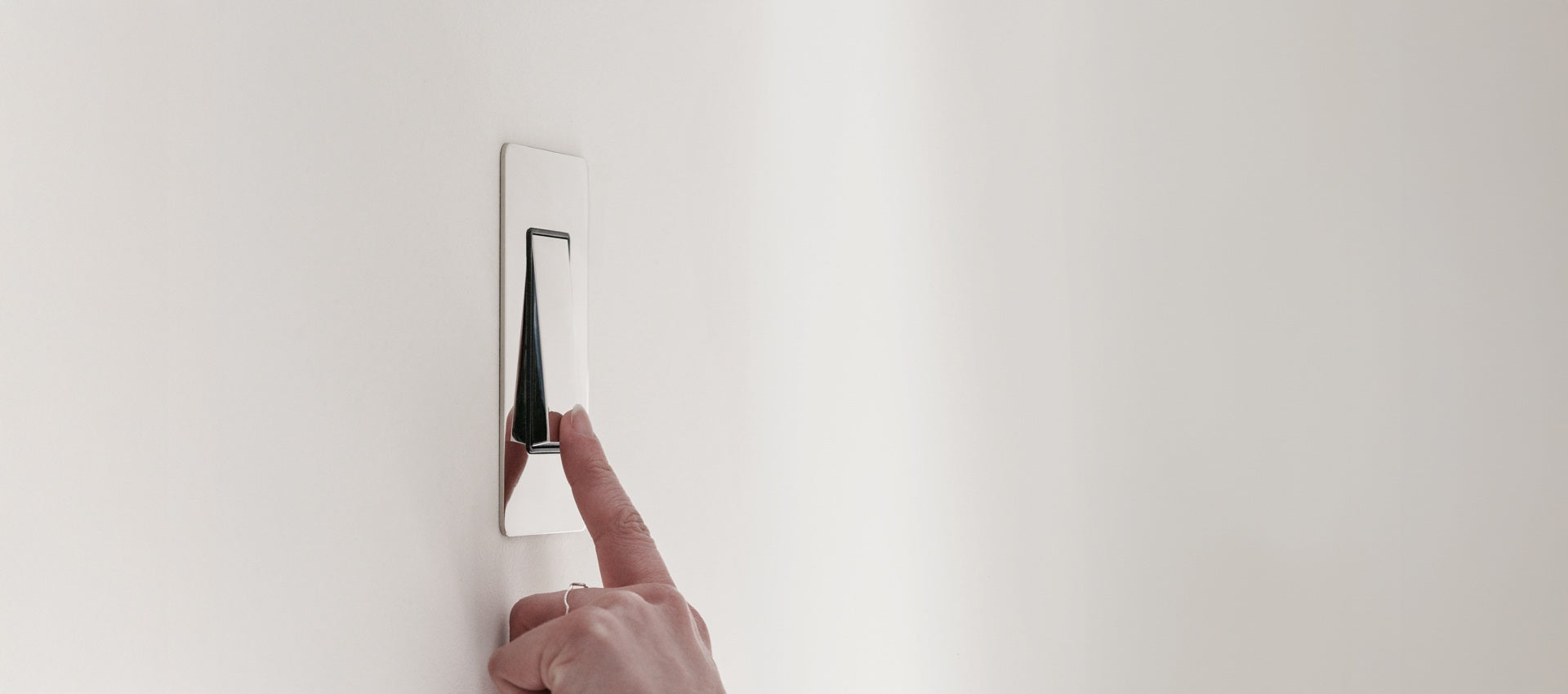 A finger pressing a sleek, modern light switch on a white wall. The switch has a polished metallic finish.
