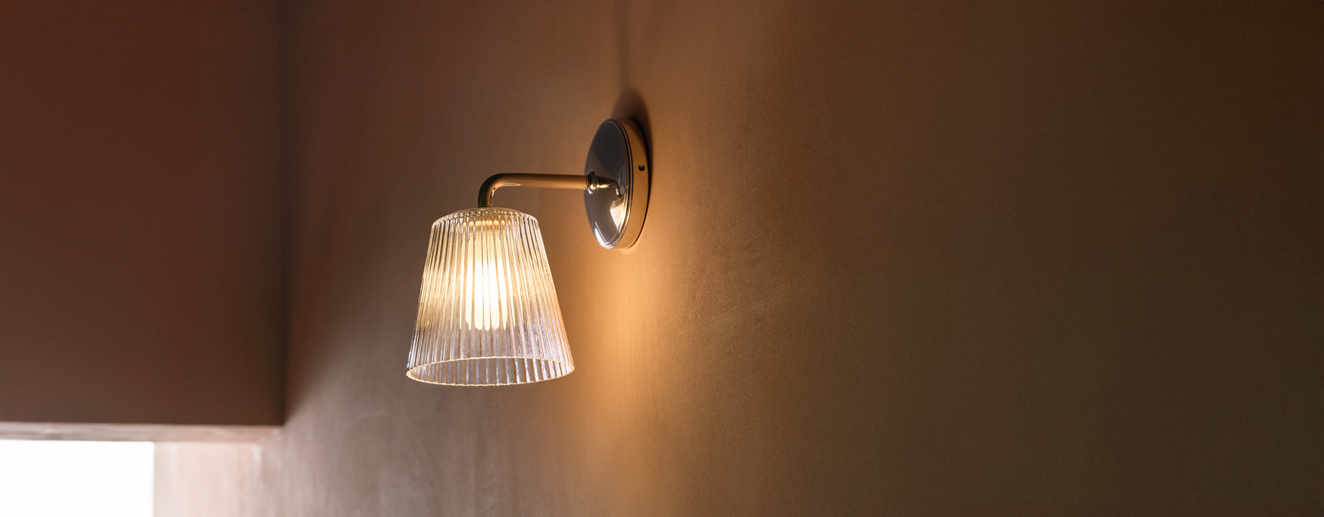 A sleek, modern wall sconce with a ribbed glass shade and warm light, mounted on a stylish bronze wall.
