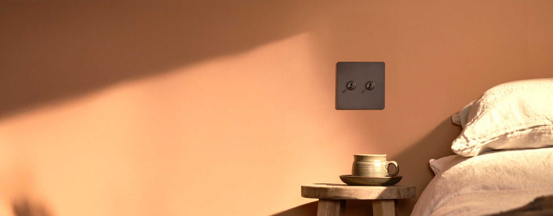 A minimalist bedside scene featuring a clay cup on a wooden table, with a wall switch above a peach-colored wall. Soft bedding is visible.

