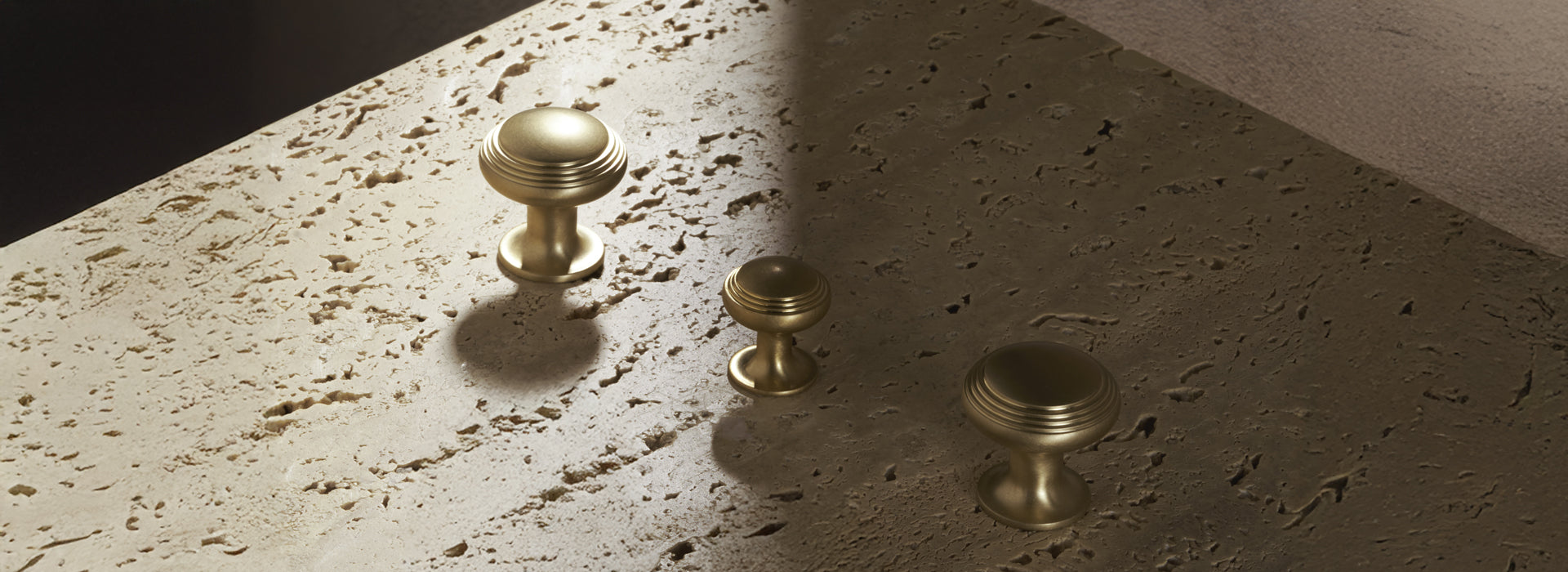 Three elegant brass knobs of varying sizes rest on a textured stone surface, illuminated by soft, natural lighting.

