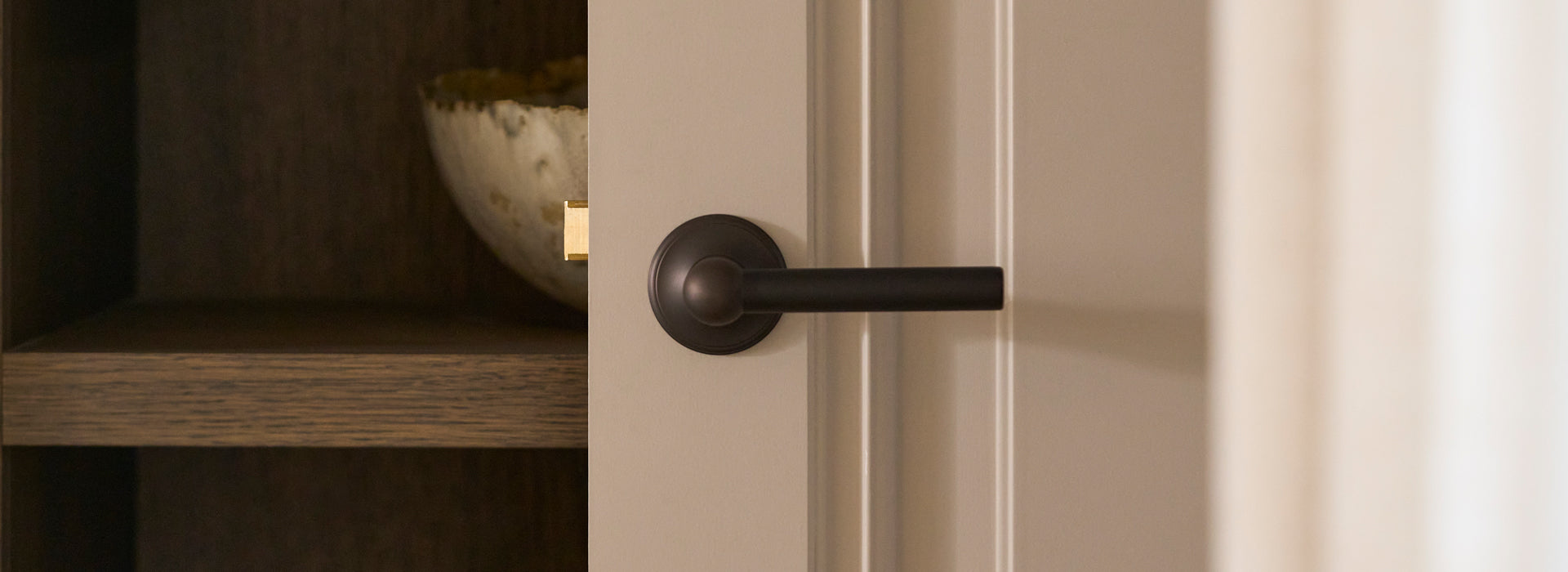 Close-up of a matte black door handle on a light-colored door, with a wooden shelf and decorative bowl partially visible behind it.
