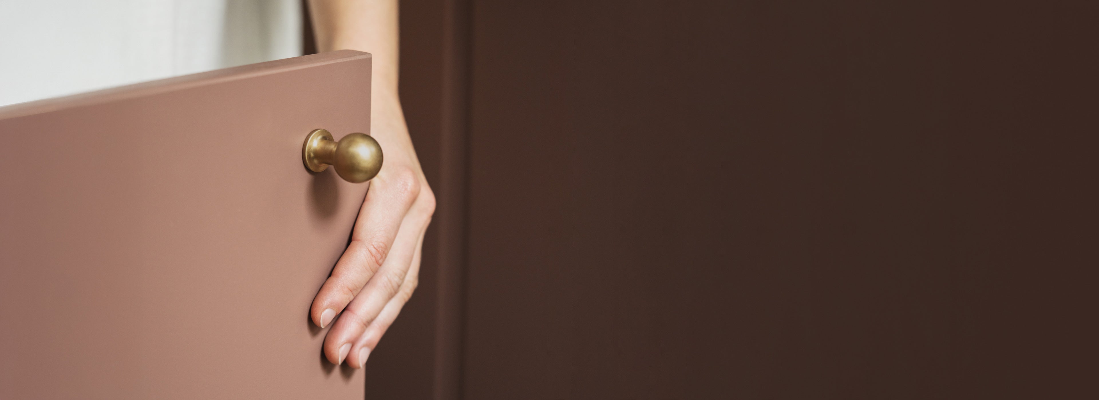 A hand gently holds a minimalist, muted pink door with a round, gold knob. Soft lighting enhances the elegant, modern design.
